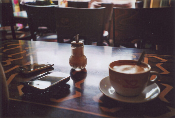 Ein Foto in warmen Brauntönen von einem Holztisch, auf dem sich eine gefüllte Cappuccinotasse, ein Zuckerstreuer sowie zwei ältere iPhones befinden.