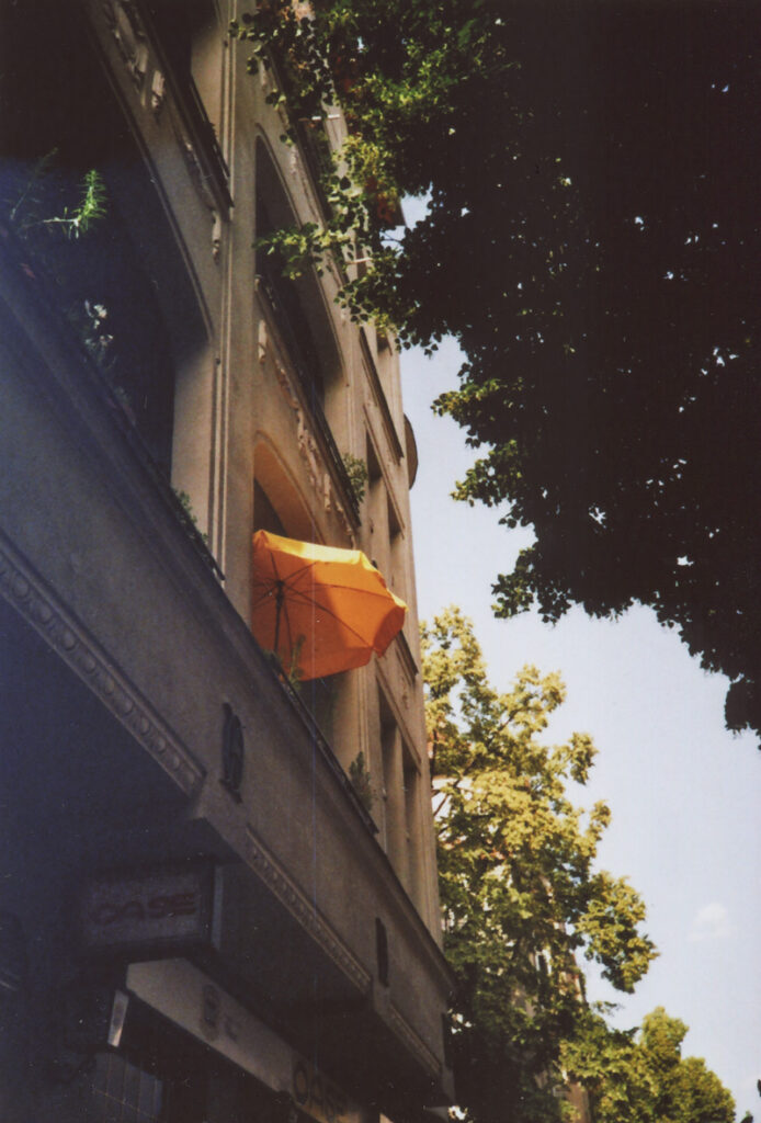 Ein orangefarbener Sonnenschirm ragt vor blauem Himmel aus einem Balkon hervor. Darunter hängt ein Schild einer Bar namens "Oase".