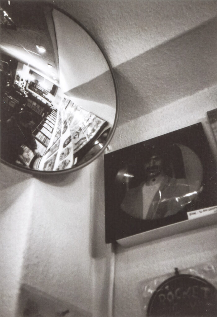 Foto von unten in die Ecke eines Plattenladens, in der ein Spiegel hängt. Darin spiegelt sich eine Person, die am Regal Platten anschaut. Darüber hängt eine LP mit dem Bild von Frank Zappa.