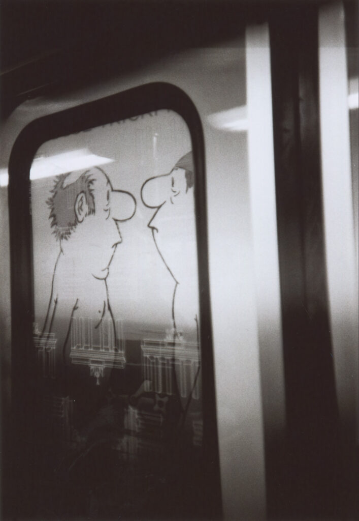 Ein schwarzweiß-Foto aus dem Tür-Fenster einer U-Bahn, an der Wand der Station ist ein Ausschnitt eines Kinoplakats für den Loriot-Film (Motiv Herren im Bad) sichtbar.