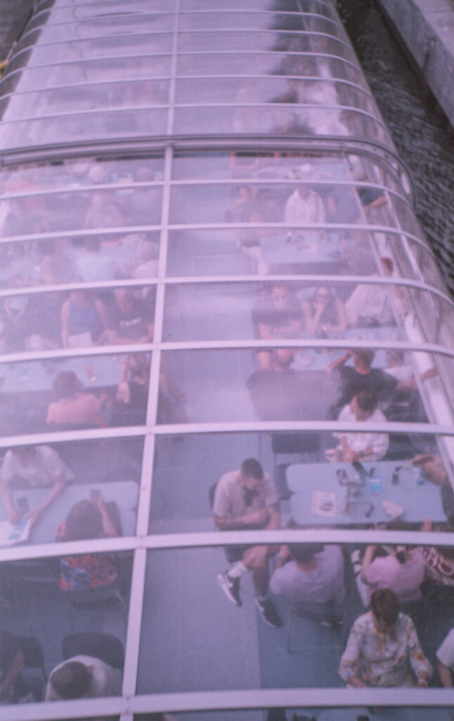 Ein analoges Foto eines Spreebootes mit einem Panoramadach aus Glas. Es ist von einer Brücke von oben fotografiert, auf dem Boot unter dem Dach sitzen Menschen an Tischen. Das Foto hat einen leichten Magenta-Stich. 