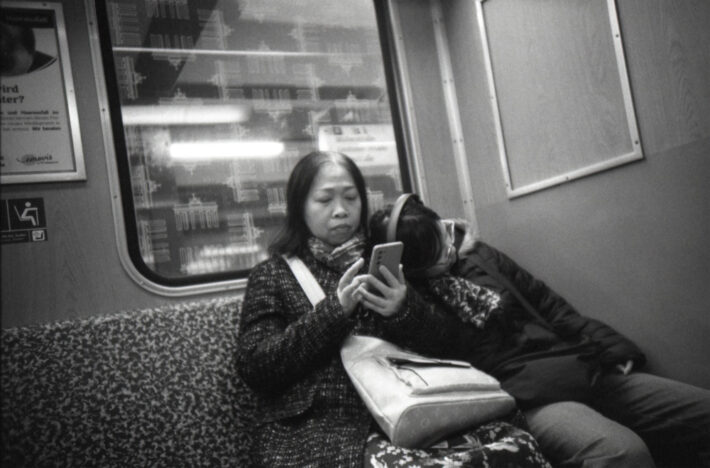 Eine Frau mittleren Alters schaut in der Berliner U-Bahn auf ihr Smartphone, eine jüngere Person, eventuell ihr Kind, liegt mit On-Ear-Kopfhörern schlafend an ihrer Schulter.