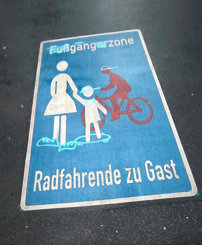 Ein auf die Straße gemaltes Schild "Fußgängerzone, Radfahrende zu Gast", aber jemand hat darüber gesprüht, so dass dort jetzt "Gangzone" steht. Die stilisierte Mutter mit Kind hat riesige Schuhe bekommen und das Kind ein Skateboard, eine Cap und eine Halskette. Die stilisierte Person auf dem Fahrrad trägt eine umgedrehte Cap. 