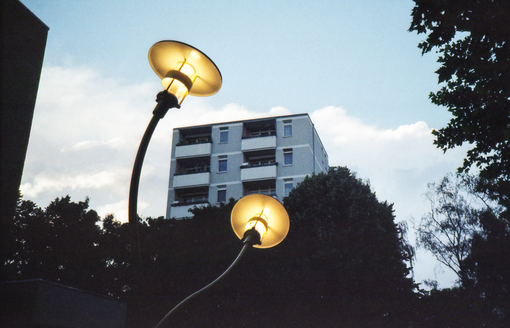 Ein frei stehendes Hochhaus in der Abenddämmerung. Im Vordergrund stehen zwei Laternen mit gebogenen Pfosten, die wirken, als seien sie aus Gummi.