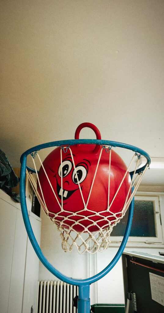 In einem Geräteraum einer Sporthalle liegt ein großer roter Hüpfball mit einem albernen Gesicht in einem Korbball-Korb. 