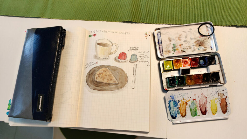 Eine Stiftetasche und ein kleiner Aquarellkasten liegen um ein Skizzenbuch herum. 
Darin ist gezeichnet eine Kaffeetasse und zwei Nespresso-Kaffeekapseln (böse, ich weiß!) und eine Piadina (ein flaches Weißbrot) auf einem Teller. 