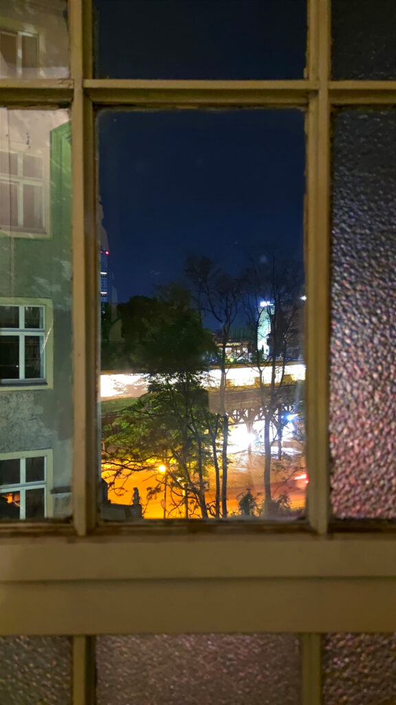 Blick aus einem kleinen Flurfenster nachts auf die Berliner U-Bahnlinie U1. Sie ist durch Bewegungsunschärfe verwischt, durch einige der kleinen Fensterscheiben zeigen sich nur bunte Lichter. 