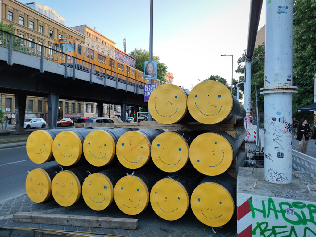 Aufeinander gestapelte sehr große und lange Röhren mit gelben geschlossenen Enden. Auf die Enden hat jemand mit Marker Smiley-Gesichter gemalt. 
Im Hintergrund fährt gerade oberirdisch eine Berliner U-Bahn vorbei (ich hatte auf eine gelbe gehofft, aber es fuhren zu der Zeit grad nur orangene). 
Ebenfalls im Hintergrund hängt ein Volt-Wahlplakat zur Europawahl auf dem "Sei kein Arschloch" steht.  