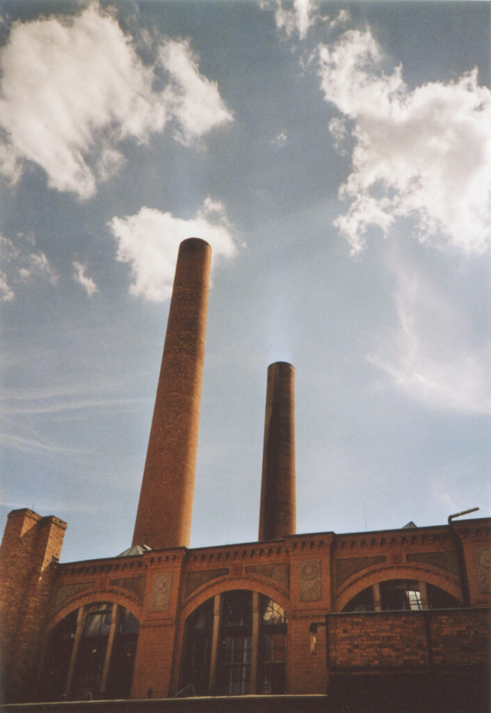 Ein rotes Backsteingebäude, eine alte Fabrik, mit zwei Schornsteinen. Mit den Wolken im blauen Himmel wirkt es, als würden die Wolken aus einem Schornstein aufsteigen.