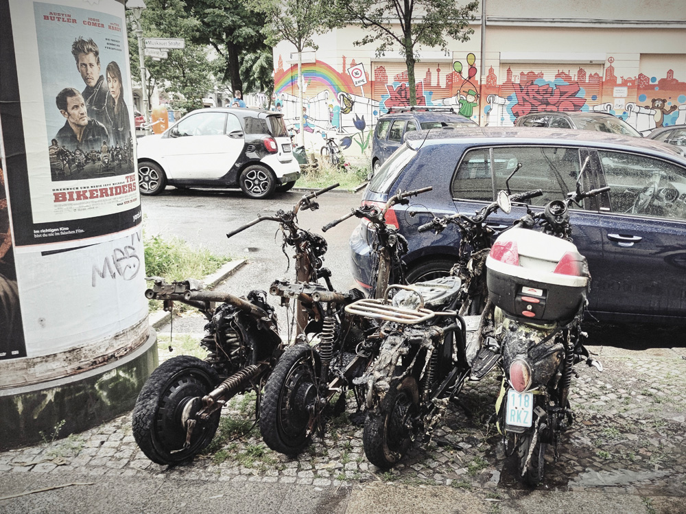 Vier ausgebrannte Roller neben einer Litfaßsäule mit einem Filmplakat von "The Bikeriders". 