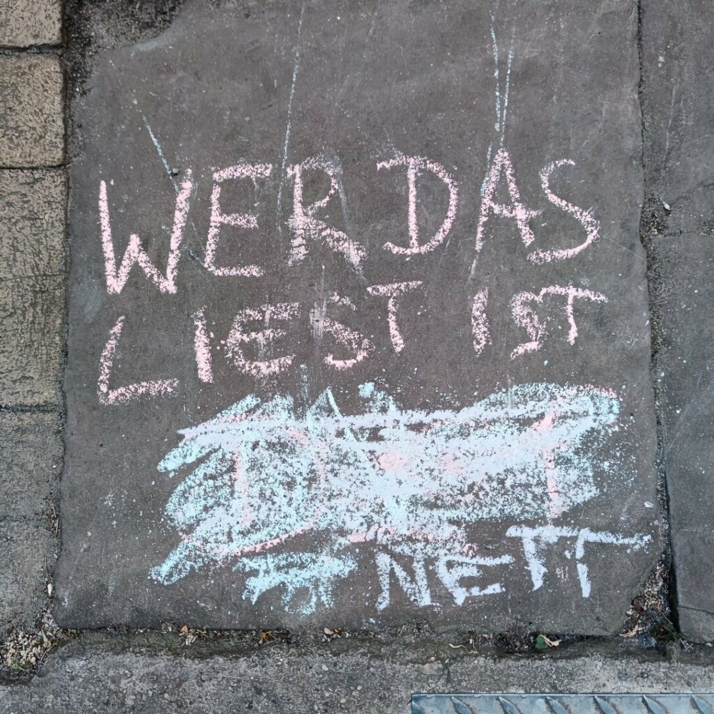 Kinder haben mit Kreide "Wer das liest ist doof" auf den Boden geschrieben. Das "doof" ist wild durchgestrichen, darunter steht "nett". 