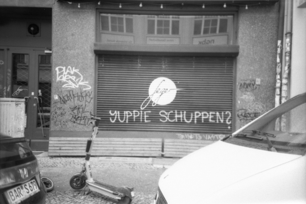 Ein analoges schwarz-weiß-Foto von einem heruntergelassenen Rollo, auf den jemand "Yuppie Schuppen?" gesprüht hat. Davor stehen Autos und ein Roller. Bei einem Auto ist das Nummernschild im Bild, dessen Buchstaben "BARS" ergibt.