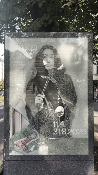Eine Werbung für eine Yoko Ono-Ausstellung. Sie hält auf dem Plakat einen Hammer in der Hand. Die Glasscheibe, hinter der das Plakat hängt, ist kaputt geschlagen.