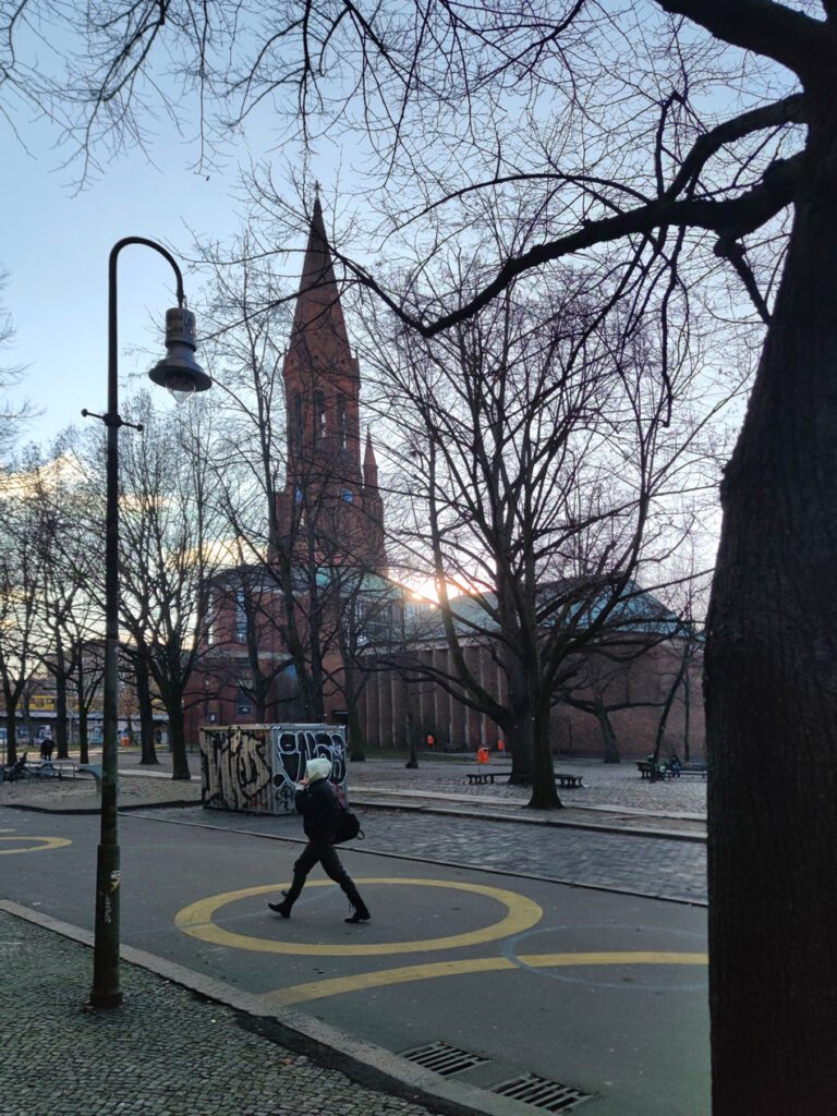 Der Lausitzer Platz in Berlin. Sonnenlicht scheint hinter einer Kirche und einem kahlen Baum hervor. 
Im Vordergrund befinden sich eine Straßenlaterne und ein weiterer Baum. Der Asphalt ist mit gelben Kreisen bemalt, in einem läuft gerade eine Person über die Straße.