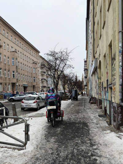 Eine Person mit schimmernder Regenbogenjacke sitzt auf einem Lastenrad gegenüber einem Amazon-Lieferfahrrad. Es wirkt, als würden beide gleich aufeinander zufahren.
