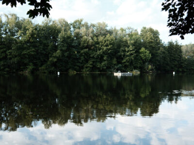 Ein See, in dem sich Bäume und Himmel spiegeln. Weiter draußen auf dem See befindet sich eine schwimmende Plattform mit einer Leiter.