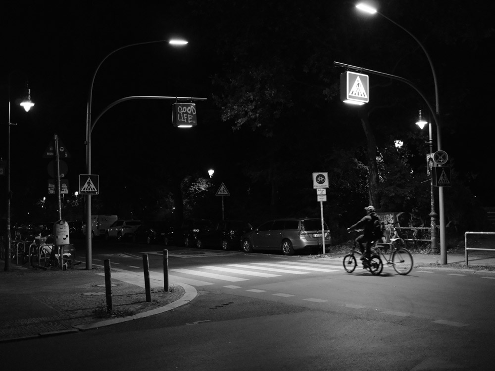 Eine Straßenkreuzung mit einem Zebrastreifen in schwarz-weiß. Auf eines Der Leuchtschilder wurde "Good Life." gesprüht. Darunter fährt gerade eine Person mit dem Fahrrad, die ein zweites Fahrrad nebenher schiebt.