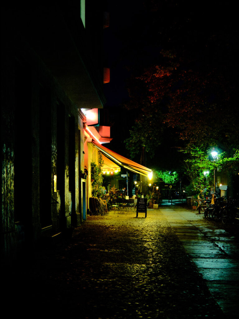 Ein Bürgersteig bei Nacht mit einem Restaurant mit einer Markise im Hintergrund. Die Kontraste von dunkel zu hell sind sehr hoch und alle Farben sehr intensiv.