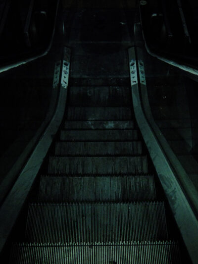 Blick von oben eine nicht mehr funktionierende Rolltreppe hinab in einem dunklen Raum. Das wenige Licht stammt von meiner Smartphone-Taschenlampe. Das Foto hat eine leicht grünliche Einfärbung und wirkt wie eine Szene aus einem Horrorfilm oder -spiel.
