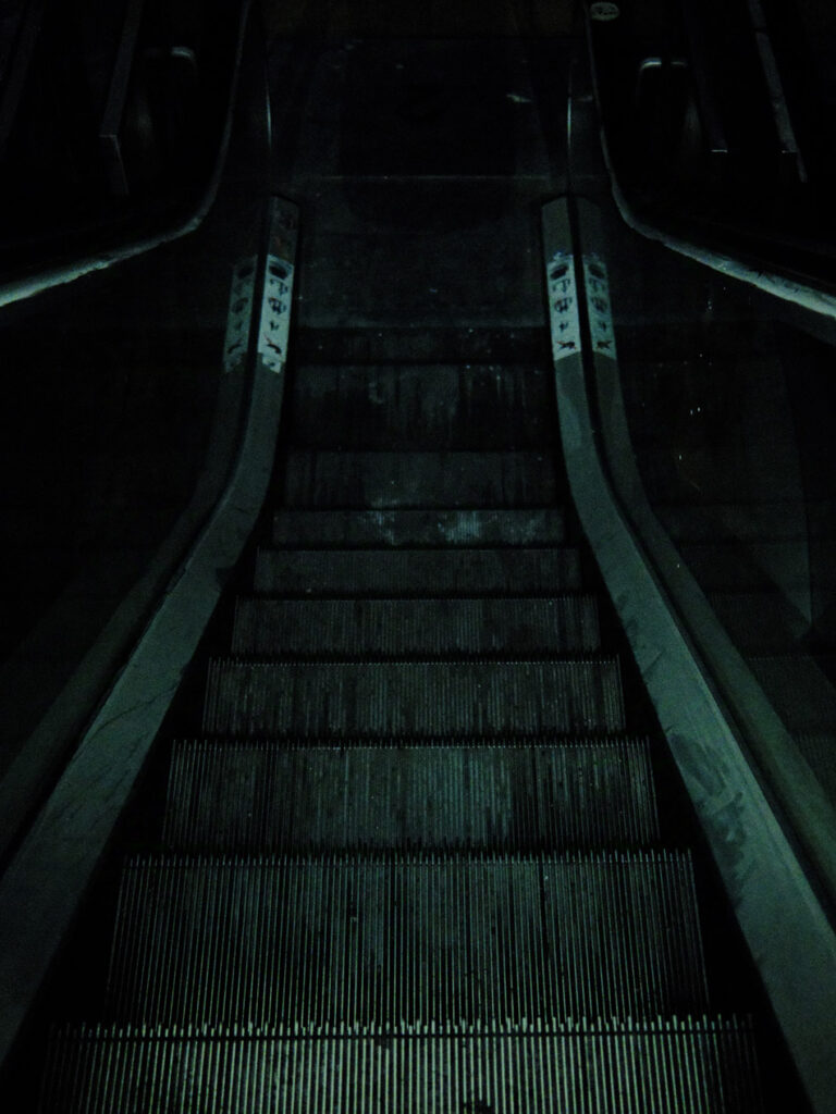 Blick von oben eine nicht mehr funktionierende Rolltreppe hinab in einem dunklen Raum. Das wenige Licht stammt von meiner Smartphone-Taschenlampe. Das Foto hat eine leicht grünliche Einfärbung und wirkt wie eine Szene aus einem Horrorfilm oder -spiel.