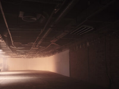 Ein schwach beleuchteter Raum, an der Decke liegen die Rohre frei. Nebel liegt in der Luft und die Töne im Bild sind in sepia gehalten.