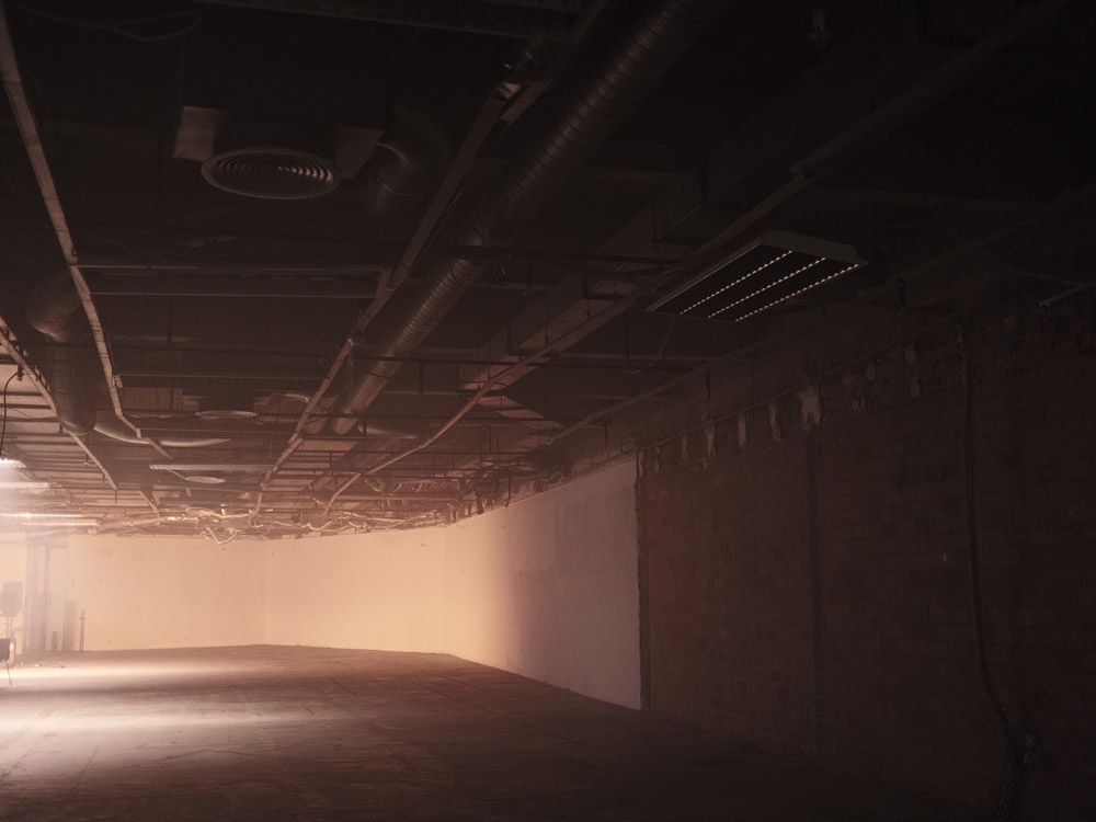 Ein schwach beleuchteter Raum, an der Decke liegen die Rohre frei. Nebel liegt in der Luft und die Töne im Bild sind in sepia gehalten.